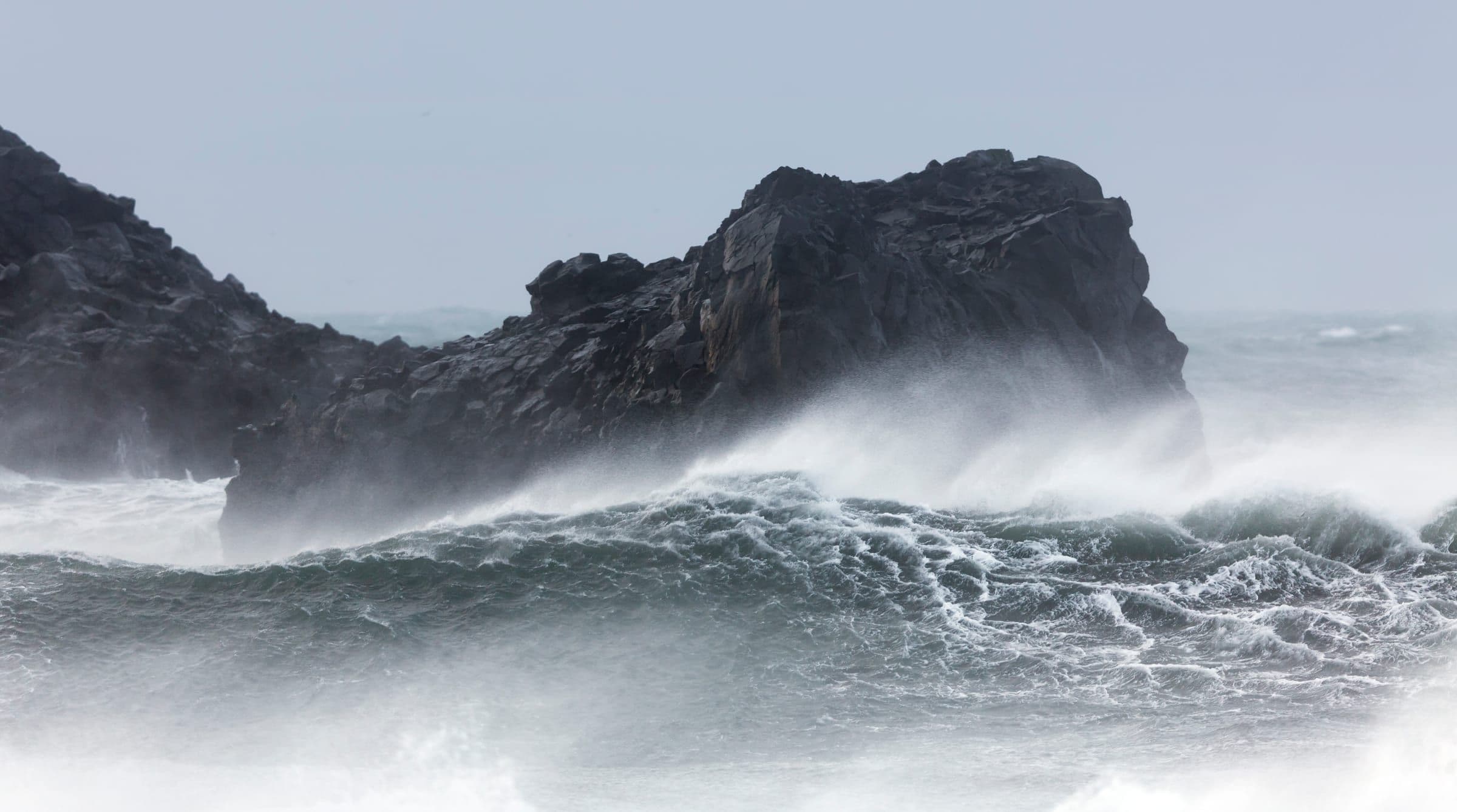 Olas del Atlántico rompiendo contra un peñasco de roca volcánica negra en la costa de Islandia, entre bruma y espuma.