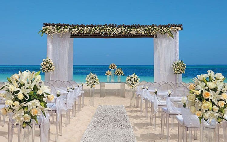 Montaje de boda en la playa: un arco de madera cubierto de flores blancas frente al mar turquesa, con sillas transparentes y un pasillo de arena.